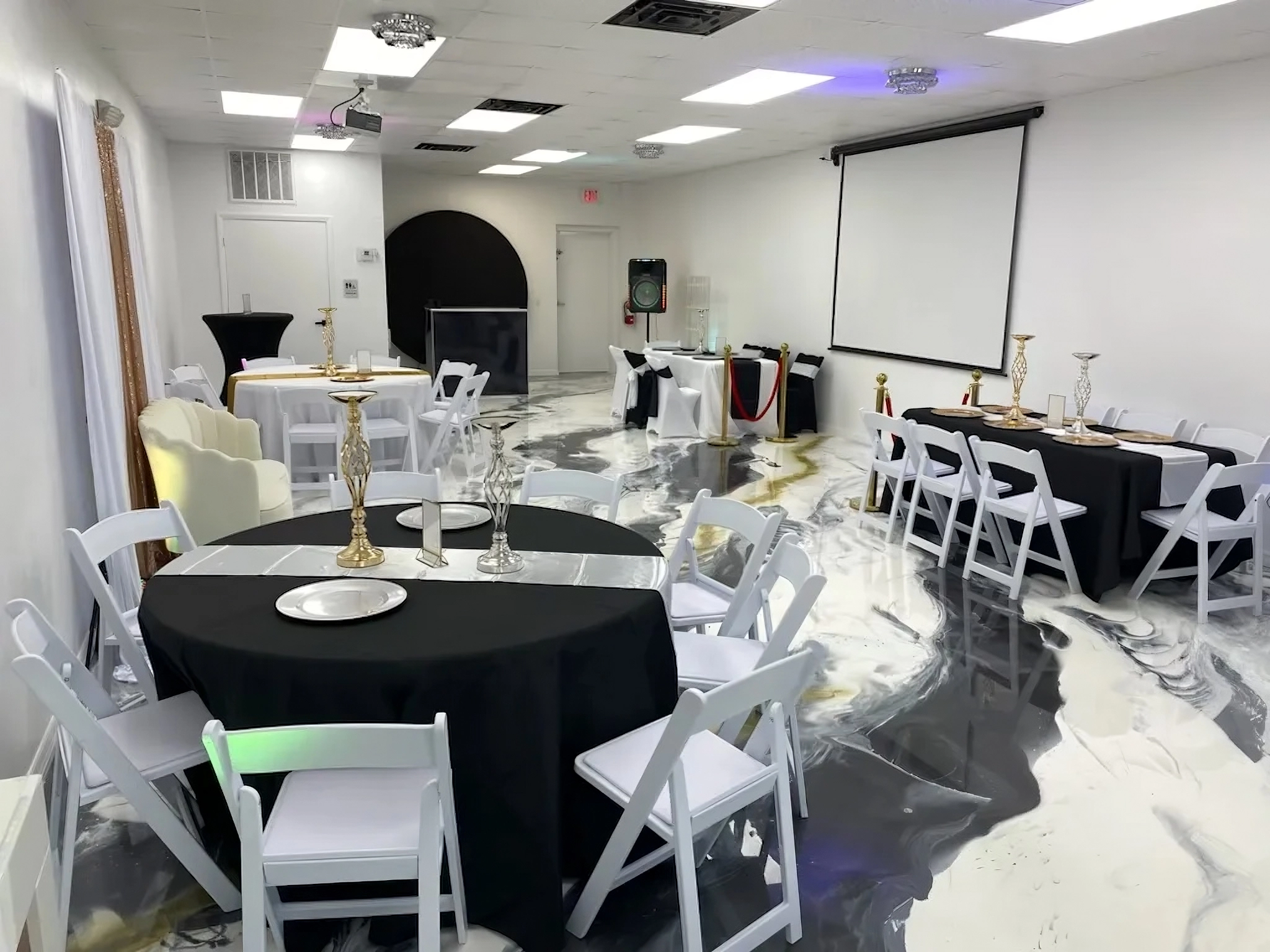 A well-lit event venue with marble-look flooring, set up for a gathering. The room features round and rectangular tables adorned with black tablecloths and decorative centerpieces. White chairs are arranged around the tables. A large screen and various seating areas are visible in the background.